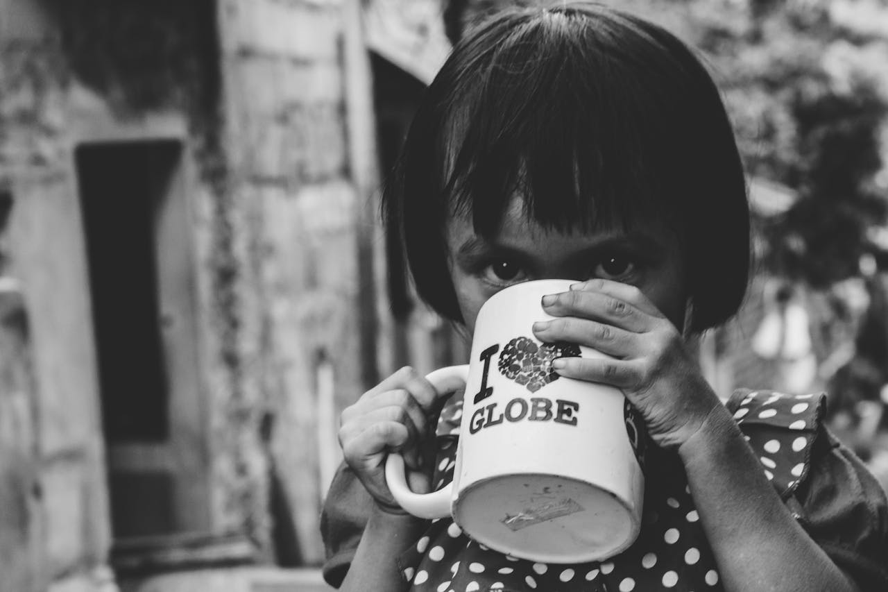 A young girl drinks from a mug in an outdoor street setting, captured in black and white.