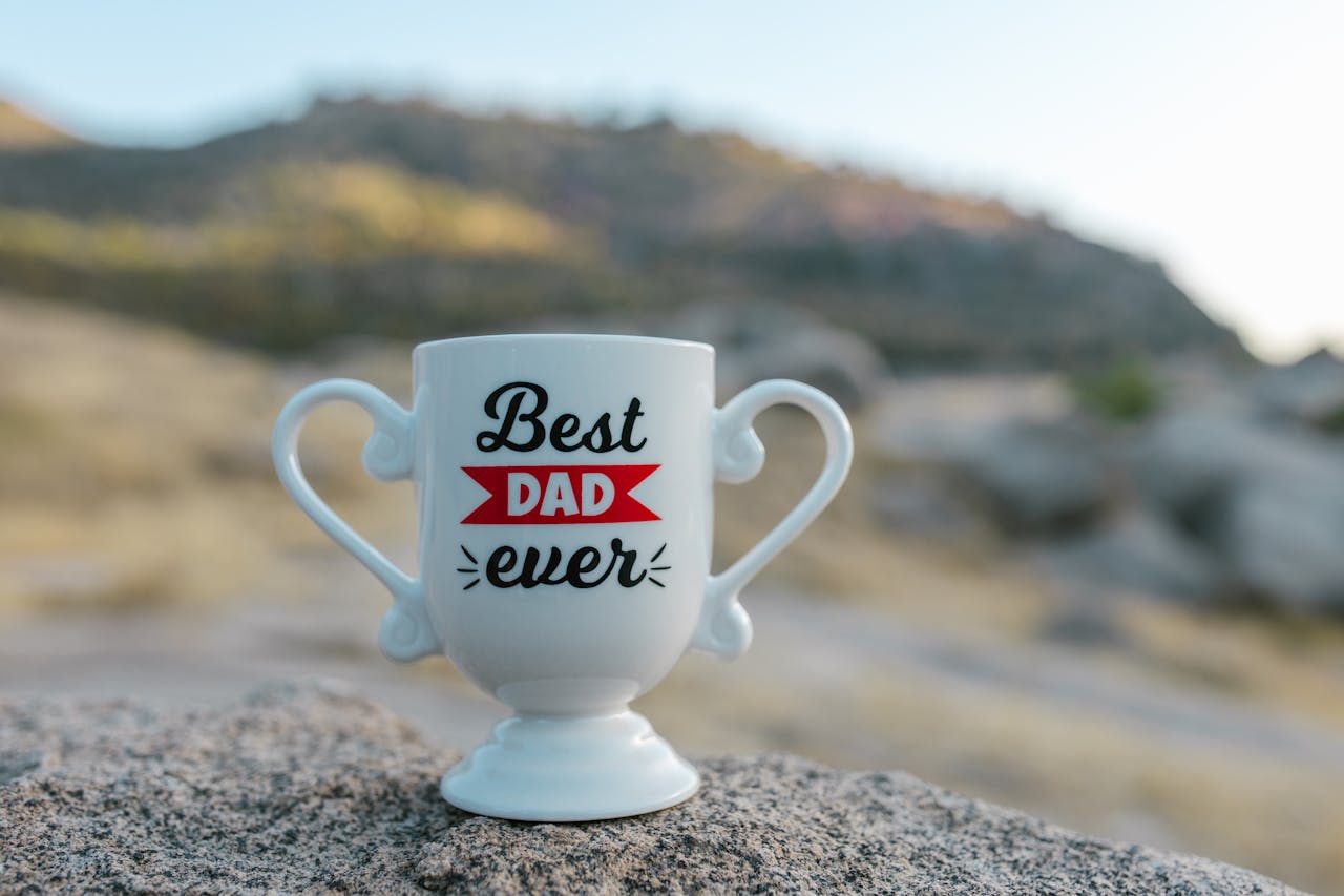 A white trophy-shaped mug with 'Best Dad Ever' text placed on a rock in a natural outdoor setting.